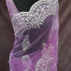 Pu&ograve; includere: Un sari lavanda con intricati ricami floreali bianchi. Il sari &egrave; drappeggiato su un manichino, mostrando il design delicato e la parola "Prisha" in basso. &Egrave; visibile anche una collana di perline bianche.