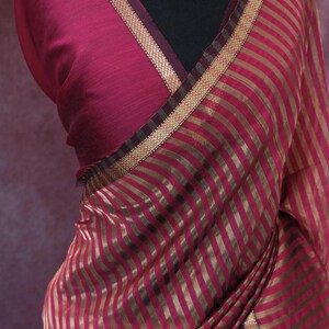 May include: A draped magenta saree with gold vertical stripes. The saree features a contrasting neckline and border with gold trim. The word "Prisha" is printed in white at the bottom of the image. A gold necklace with red and white accents is visible.