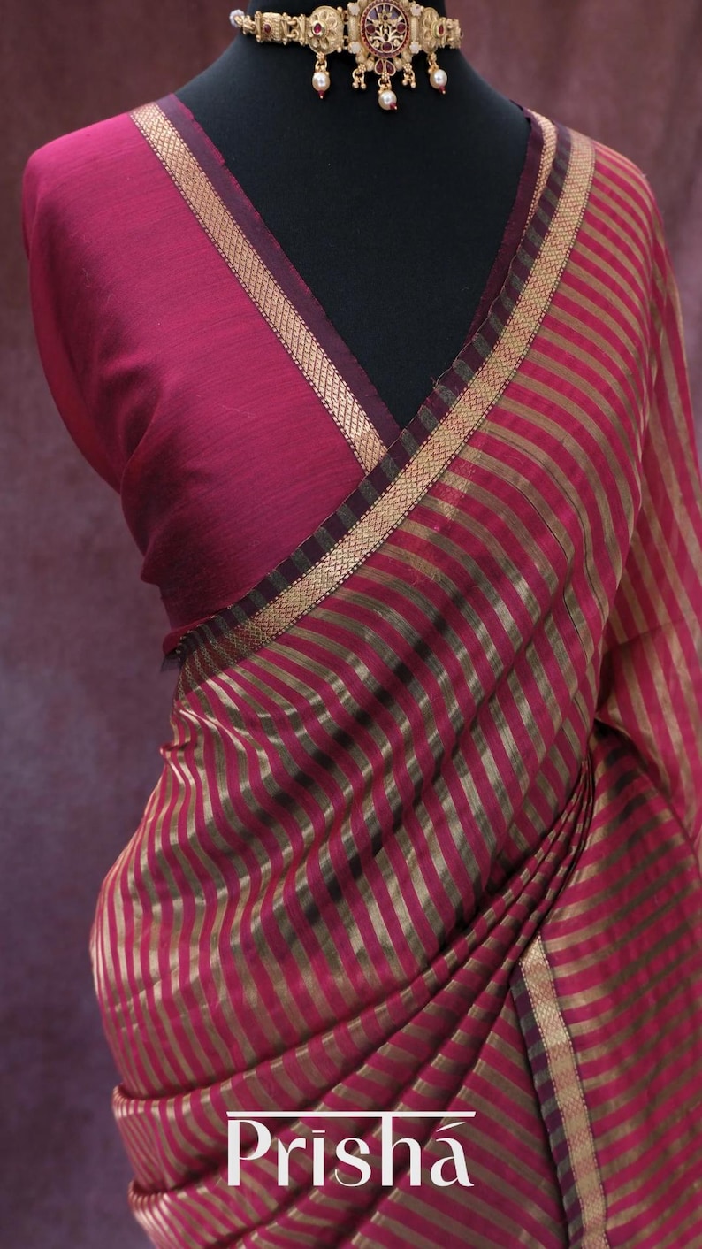 May include: A draped magenta saree with gold vertical stripes. The saree features a contrasting neckline and border with gold trim. The word "Prisha" is printed in white at the bottom of the image. A gold necklace with red and white accents is visible.