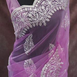 Pu&ograve; includere: Un sari lilla trasparente con intricati ricami floreali bianchi e un bordo abbinato. Il sari &egrave; drappeggiato su un manichino, mettendo in risalto il tessuto delicato e il design. La parola "Prisha" &egrave; visibile in basso.