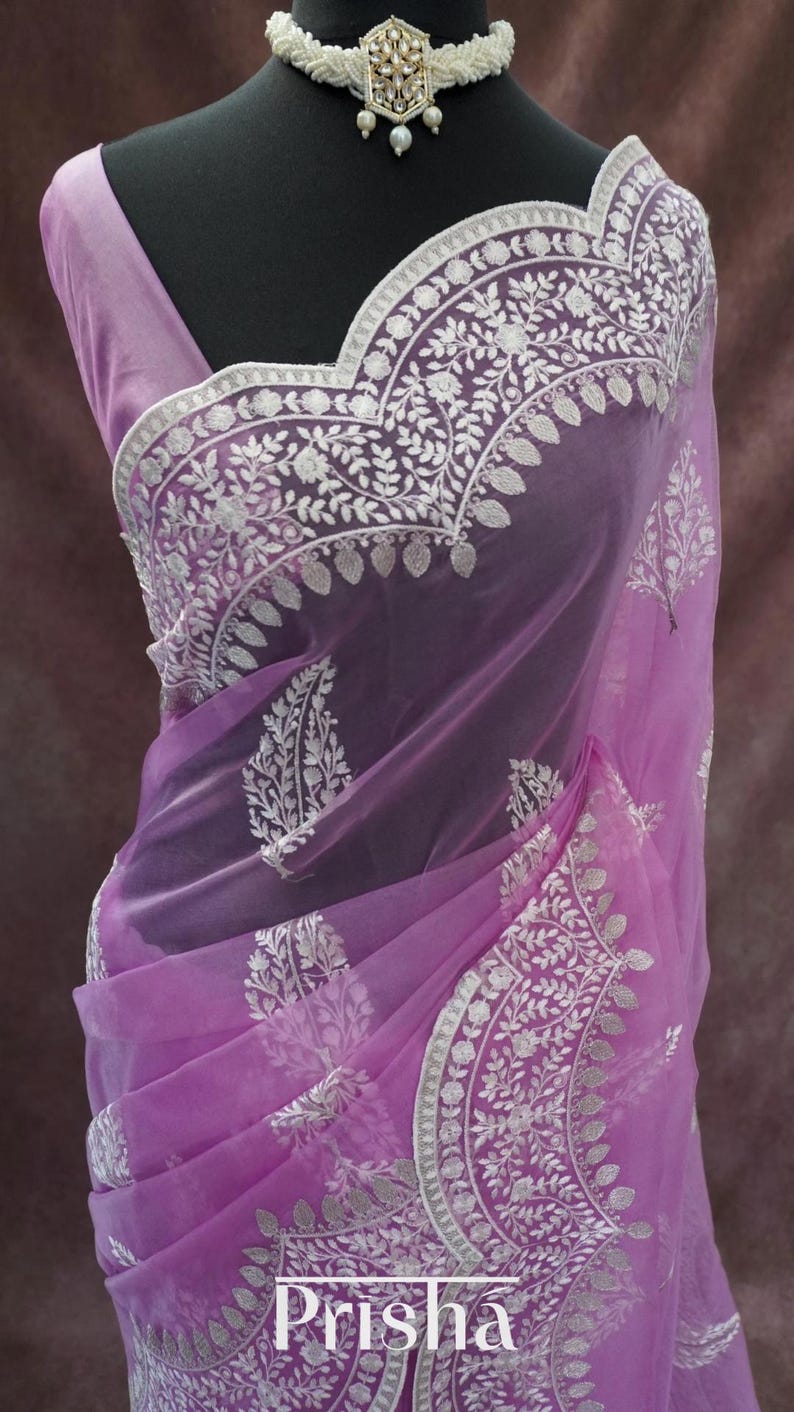 Pu&ograve; includere: Un sari lilla trasparente con intricati ricami floreali bianchi e un bordo abbinato. Il sari &egrave; drappeggiato su un manichino, mettendo in risalto il tessuto delicato e il design. La parola "Prisha" &egrave; visibile in basso.