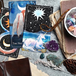 Op de afbeelding: Vier tarotkaarten, De Wereld, De Hogepriesteres, De Ster en XVII De Ster, liggen uitgespreid op een tafel met een kom met kleurrijke kristallen, een boek en een kralenketting.