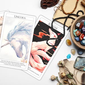 May include: Three tarot cards with watercolor illustrations of a Gorgon, a Unicorn, and a Kitsune. The cards are laid out on a white surface with various crystals, a wooden bowl, and a feather.