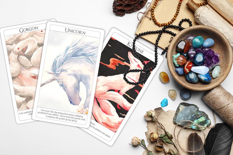 May include: Three tarot cards with watercolor illustrations of a Gorgon, a Unicorn, and a Kitsune. The cards are laid out on a white surface with various crystals, a wooden bowl, and a feather.