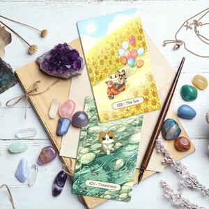 Puede incluir: Vista cenital de una colecci&oacute;n de cartas del tarot, cristales y objetos vintage. Se ven dos cartas del tarot, una que representa un gato en un scooter con globos, la otra un gato en el agua. Varios cristales en tonos morados, rosas, azules y verdes est&aacute;n dispersos.