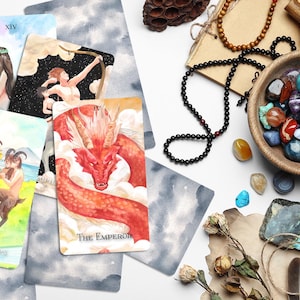 Op de afbeelding: Een spreiding van tarotkaarten op een wit oppervlak met kristallen, een houten kom en een veer. De kaarten omvatten De Keizer, XIV en Page of Cups.