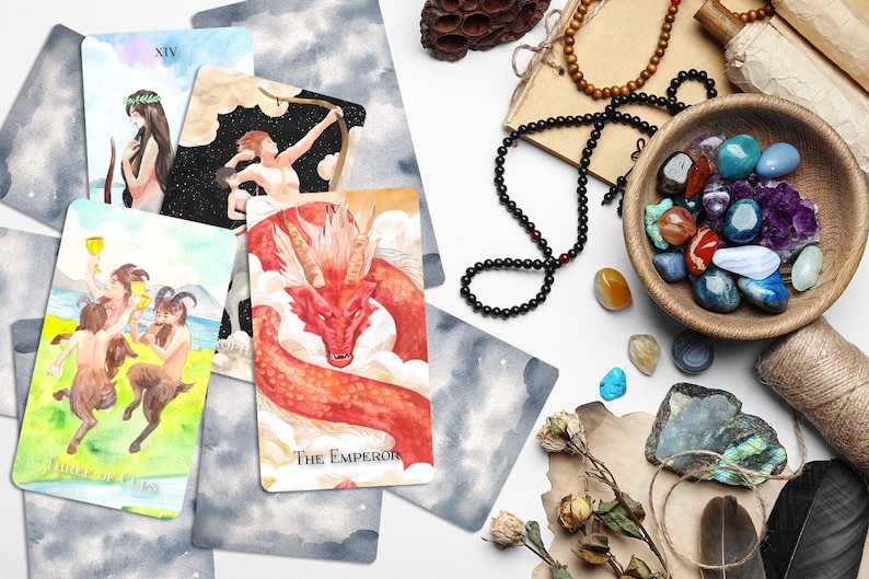 Op de afbeelding: Een spreiding van tarotkaarten op een wit oppervlak met kristallen, een houten kom en een veer. De kaarten omvatten De Keizer, XIV en Page of Cups.