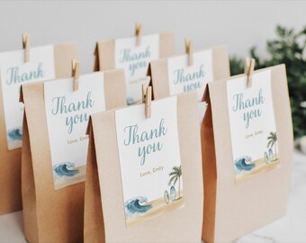 May include: Kraft paper gift bags with white tags that read "Thank you" in teal script. Each tag features a beach scene with waves, palm trees, and surfboards. The tags are held in place with small wooden clothespins.