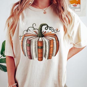 May include: Cream-colored t-shirt featuring a pumpkin design made of books. The pumpkin has a green stem and vine details. The books are in shades of orange, brown, and red, with visible pages. A fall-themed graphic tee.