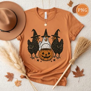 May include: Orange t-shirt featuring a Halloween design. The graphic shows three chickens wearing witch hats, standing around a carved pumpkin. The design is in black and white, with a few stars below the pumpkin.