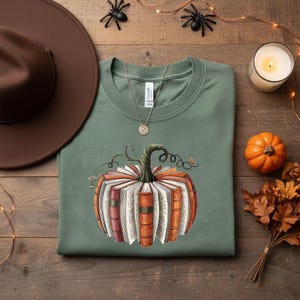 May include: Sage green t-shirt featuring a pumpkin design made of books. The pumpkin has a green stem and is surrounded by orange and white book pages. A brown hat, candle, and small pumpkin are on a wooden surface.