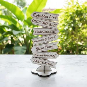 May include: A white wooden signpost with multiple directional signs. Each sign has a brown border and text such as "Forbidden Love" and "Only One Bed". The signpost is on a white surface with a blurred green background.