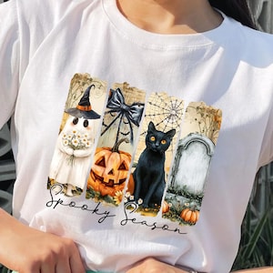 May include: White t-shirt featuring a Halloween-themed design. The design includes a ghost holding flowers, a jack-o'-lantern, a black cat, and a tombstone. The words "Spooky Season" are written in a script font below the images.