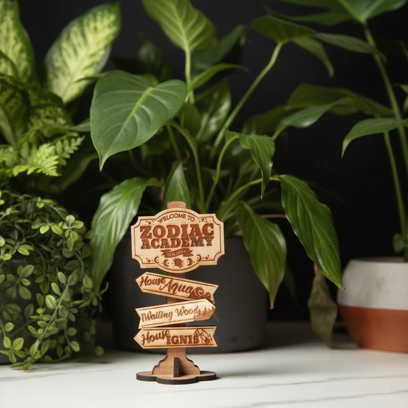 Op de afbeelding: Een houten bord met de tekst "Welcome to Zodiac Academy" en pijlen voor "House Aqua", "Wailing Woods" en "House Ignis". Het bord is lichtbruin en staat voor weelderige groene planten.