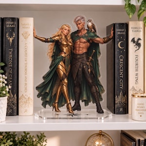May include: A clear acrylic standee featuring a fantasy duo: a blonde elf in gold armor and a green cape, and a shirtless man with silver hair, a green cape, and a small bird on his shoulder, standing on a hexagonal base between two books.
