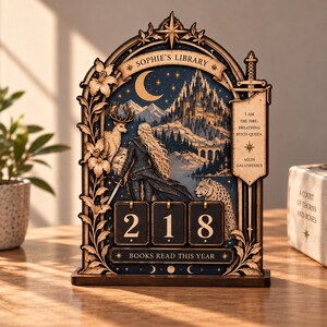 Könnte beinhalten: Ein hölzerner, bogenförmiger Tischkalender mit dem Text SOPHIE'S LIBRARY und BOOKS READ THIS YEAR. Er zeigt eine Fantasy-Illustration mit einer Figur, einem Schwert, einem Hirsch, einem Leoparden und einer Burg. Ein Banner trägt den Text I AM THE FIRE-BREATHING BITCH-QUEEN.