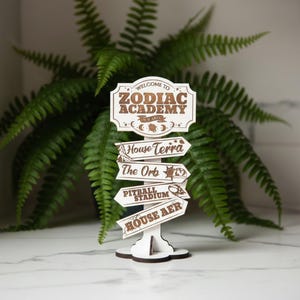 Op de afbeelding: Een houten wegwijzer met de tekst "Welcome to Zodiac Academy" en pijlen die wijzen naar "House Terra", "The Orb", "Pitball Stadium" en "House Aer". Het bord is wit met bruine letters en decoratieve elementen.