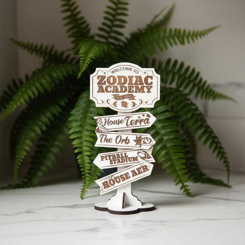 Op de afbeelding: Een houten wegwijzer met de tekst "Welcome to Zodiac Academy" en pijlen die wijzen naar "House Terra", "The Orb", "Pitball Stadium" en "House Aer". Het bord is wit met bruine letters en decoratieve elementen.