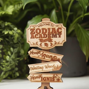Op de afbeelding: Houtkleurige wegwijzer met de tekst "Welcome to Zodiac Academy Solaria". Onder de wegwijzer staan drie pijlen met de tekst "House Aqua", "Wailing Wood" en "House Ignis".