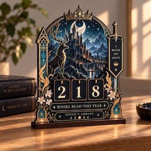 Puede incluir: Un expositor decorativo de madera con un paisaje de fantasía que incluye un ciervo, un castillo y una luna creciente. Incluye tres bloques numéricos giratorios que muestran 218, con el texto A BRAVER YOU AWAITS, BOOKS READ THIS YEAR y SOPHIE'S LIBRARY.