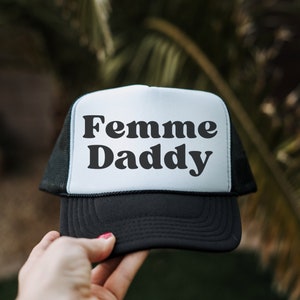 Op de afbeelding: Een zwart-witte truckercap met de tekst "Femme Daddy" in een retro-lettertype.