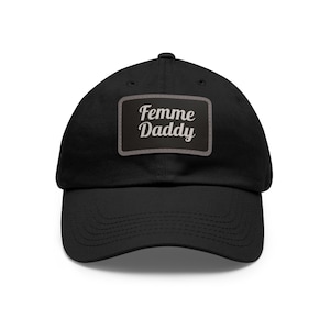 Può includere: Un cappello da baseball nero con una toppa grigia che dice "Femme Daddy" in un font bianco corsivo.