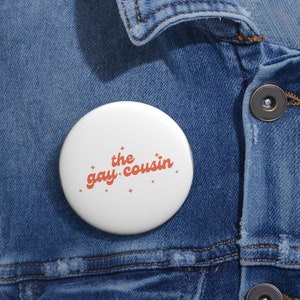 May include: White button with the text "the gay cousin" in orange with small white stars around the text.