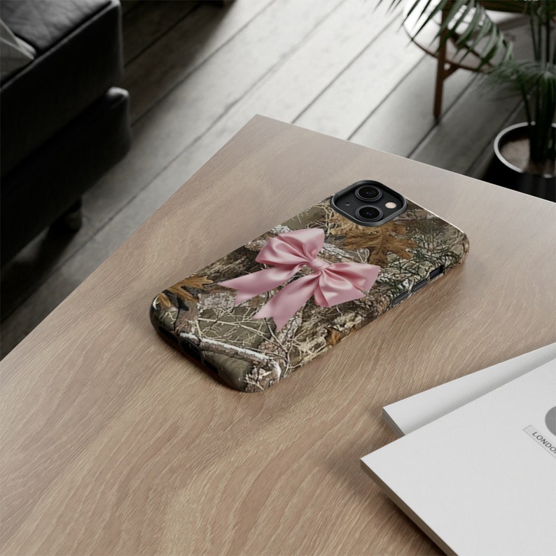 Pode incluir: Uma capa de telefone camuflada com um la&ccedil;o rosa. A capa &eacute; projetada para um smartphone com uma protuber&acirc;ncia da c&acirc;mera.