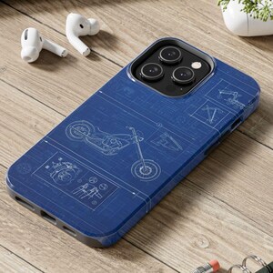 May include: A blue phone case with a motorcycle blueprint design. The case has a black border and a triple-lens camera cutout. White earbuds are visible to the left of the phone case.