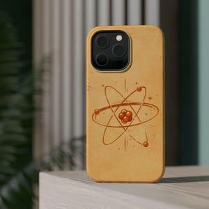 May include: A tan phone case with a retro atom design in brown and orange. The case has a rounded shape and a camera cutout. The background is blurred, suggesting an outdoor setting.