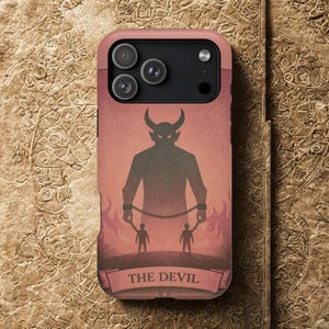 May include: A phone case with a devil-themed design. The case has a pink border and a graphic of a horned silhouette with chains, two figures, and the text "THE DEVIL" on a banner. The background is a reddish-brown.