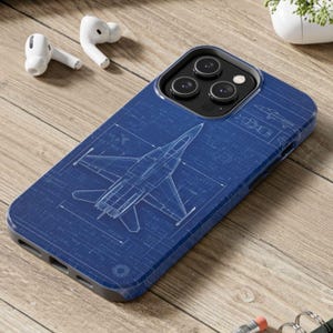 May include: A blue phone case featuring a detailed blueprint design of a fighter jet. The case has a dark border and a glossy finish. White wireless earbuds are visible in the background, along with a small white vase with greenery.