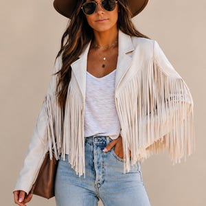 Puede incluir: Una chaqueta de color crema con largos flecos en el pecho y las mangas, combinada con una camiseta blanca de cuello en V, vaqueros azul claro, un sombrero marrón de ala ancha, gafas de sol oscuras y un bolso de mano marrón.