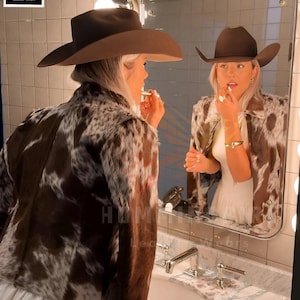 May include: A brown cowboy hat and a cowhide jacket with brown and white patterns are reflected in a mirror. The image also shows a white dress and a bathroom sink. The logo for Hummingbird Leather Wears is visible.