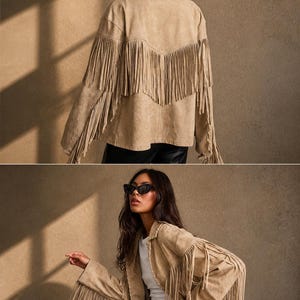 K&ouml;nnte beinhalten: Beigefarbene Wildlederjacke mit langen Fransen an den &Auml;rmeln und am R&uuml;cken. Die Jacke hat einen klassischen Western-Stil mit Kragen und lockerer Passform. Das Model tr&auml;gt eine schwarze Sonnenbrille und eine schwarze Hose.