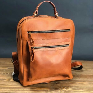 May include: A brown leather backpack with a top handle and multiple zippered pockets. The backpack has a rectangular shape with a rounded top and a handle at the top. The zippers are black, and the leather has a smooth texture.