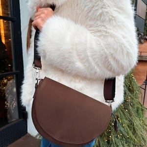 May include: A brown half-moon shaped leather crossbody bag with a black strap. The bag is worn by a person wearing a white faux fur coat and blue jeans.