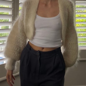May include: A white fuzzy cropped cardigan with long sleeves and a slightly open front. The cardigan is worn over a white tank top and black pants.