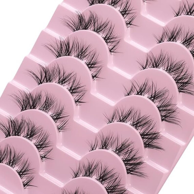 K&ouml;nnte beinhalten: Ein Set mit zehn einzelnen falschen Wimpern auf einer rosa Plastikpalette. Die Wimpern sind in Reihen angeordnet, wobei jede Reihe einen anderen Wimpernstil enth&auml;lt.