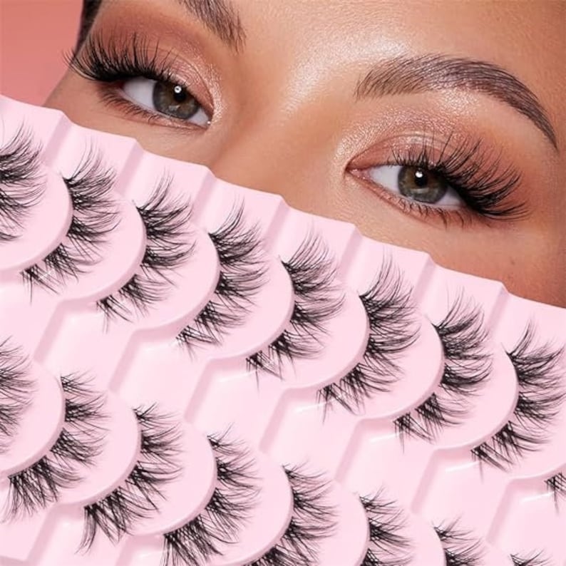 K&ouml;nnte beinhalten: Nahaufnahme einer pinkfarbenen und wei&szlig;en Packung mit falschen Wimpern. Die Wimpern sind in Reihen angeordnet, wobei jede Reihe einen anderen Wimpernstil enth&auml;lt. Die Wimpern bestehen aus synthetischen Fasern und sind f&uuml;r die Anwendung auf der oberen Wimpernlinie konzipiert.