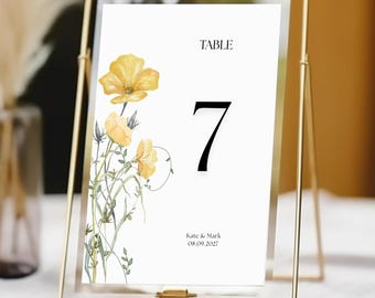 May include: A white table number card with a yellow floral design and the number 7 printed in black. The card is in a gold metal frame and reads "Table 7" with the text "Kate & Mark 08.09.2027" at the bottom.