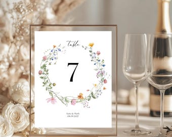 May include: A white table number card with a floral wreath design in shades of pink, blue, and yellow. The card reads "Table 7" and "Kate & Mark 08.09.2022".