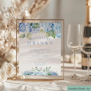 May include: A framed sign with the words "Signature Drinks" in blue script, featuring a floral design of blue hydrangeas. The sign lists two cocktail recipes: Blueberry Bash Smash and Honeydew Mojito. The names "Kate & Mark" and the date August 20, 2028 are also displayed.