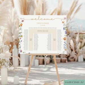 以下が含まれることがあります： 結婚式のウェルカムボード。名前「EMILY & MARK」と日付「07.10.28」が記載されています。木製のイーゼルに飾られ、花柄の装飾が施されています。座席表が含まれています。