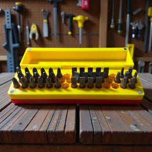 May include: A yellow and red tool organizer with a variety of dark gray metal drill bits. The organizer is open, revealing the bits arranged in a grid pattern. The background shows tools hanging on a pegboard.