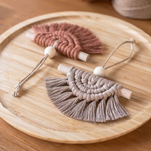 Puede incluir: Dos colgantes de pared de macramé en tonos marrón y beige, cada uno con una clavija de madera y una cuenta de madera. Están dispuestos sobre una bandeja redonda de madera. Las piezas de macramé tienen un diseño en forma de abanico.