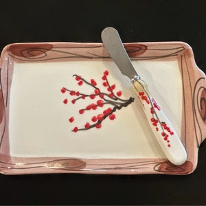 May include: A white ceramic serving tray with a pink border and a painted design of a red berry branch. A silver butter knife with a white handle and a red berry design is on the tray.