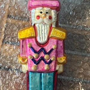 May include: A pink and gold glass Christmas ornament shaped like a nutcracker soldier. The ornament has a green hat with gold trim and a white face with a black mustache and beard.