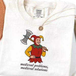 May include: Ivory-colored t-shirt featuring a cartoon jester holding an axe. The jester wears a red and yellow outfit. The text "medieval problems, medieval solutions" is printed below the graphic. The shirt has a Comfort Colors tag.
