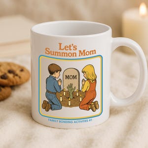 May include: White ceramic mug with the text "Let's Summon Mom" and an illustration of two children kneeling before a tombstone. The tombstone reads "MOM" and a zombie hand is reaching out. The mug also has the text "Family Bonding Activities #1".